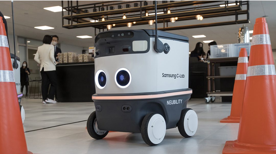 An autonomous robot labeled “Samsung C-Lab” and “NEUBILITY” moves indoors between orange safety cones. People are gathered in the background, suggesting a startup showcase or demo event.